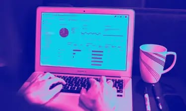 Person typing on a laptop displaying data analytics charts next to a coffee mug in purple light.