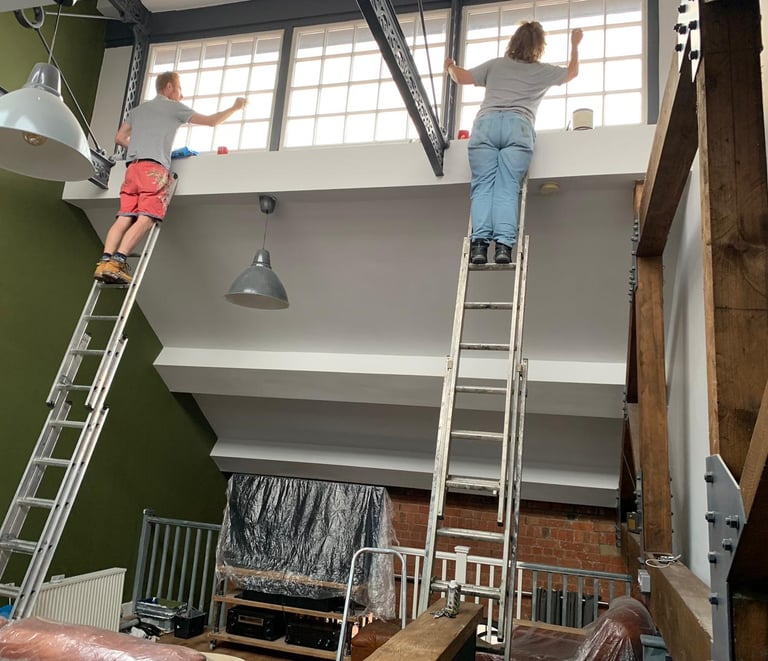 An in-progress living room, with a slanted ceiling and windows, and two painters working on ladders.