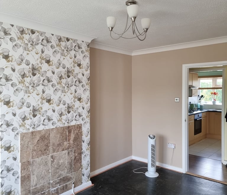An unfurnished living room, with pink/cream walls, except a wall with white, floral wallpaper.
