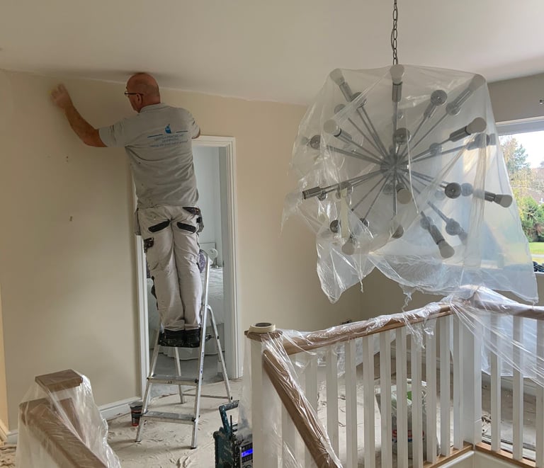 An in-process white landing/hallway, with sheet covered furniture and a painter on a ladder.