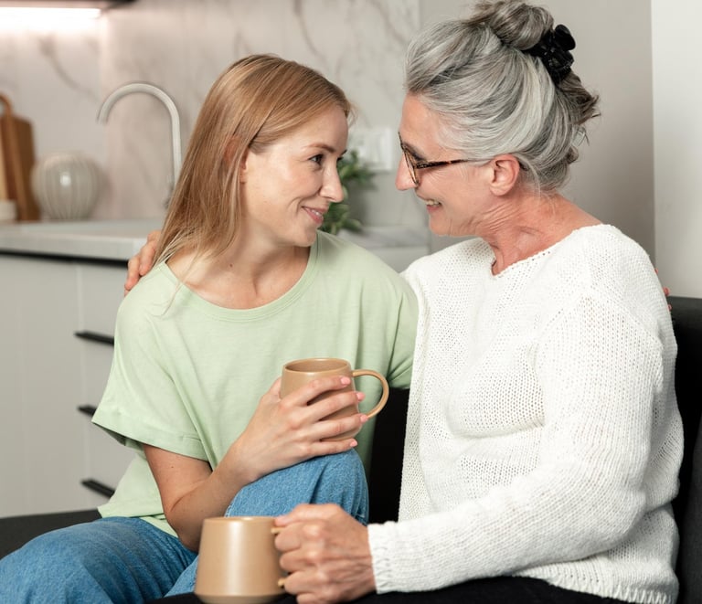 A smiling senior mother and adult daughter talking while holding coffee mugs on a sofa at home.