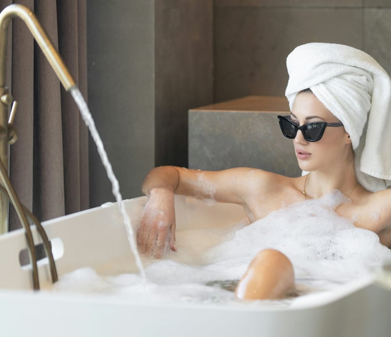 woman wearing glasses and hair wrapped in towel, relaxing in modern tub filled with water and bubbles.