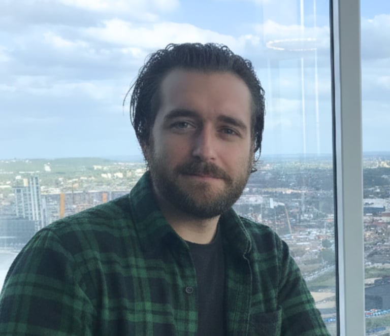 Professional man in a green flannel shirt posing by a window with a city skyline view.