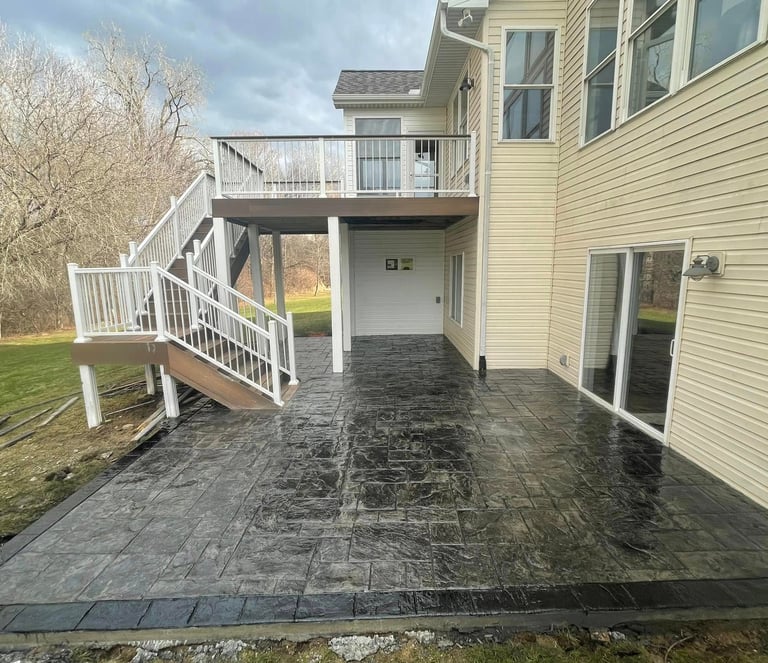 New dark grey stamped concrete patio installed under a second-story wooden deck with white railings.