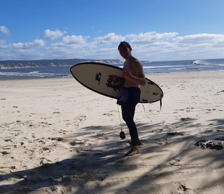 Surf coach Ben standing on a tropical beach holding his surfboard and giving a shaka.