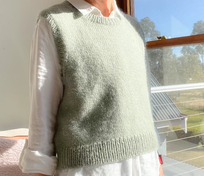 Woman wearing a dusty green hand knit slipover with a white shirt and jeans in a sunny room in front of a window. 