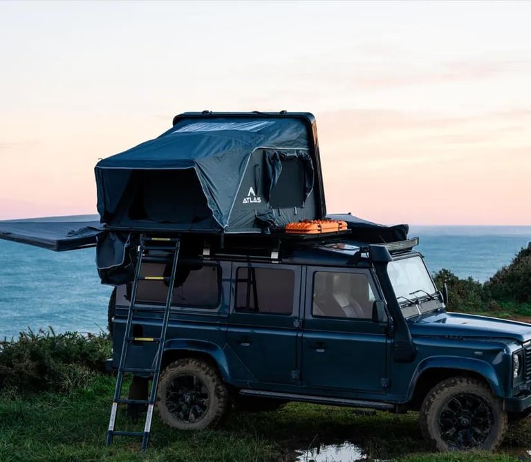 Land Rover Defender with a rooftop tent and awning parked on a scenic coastal cliff at sunset.