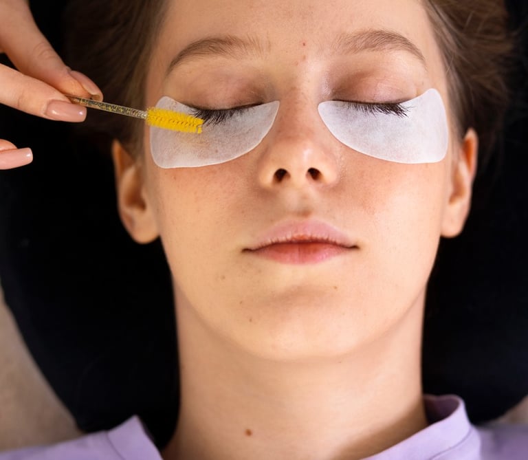 A beauty technician uses a yellow spoolie brush to groom eyelashes during a professional eyelash extension treatment.