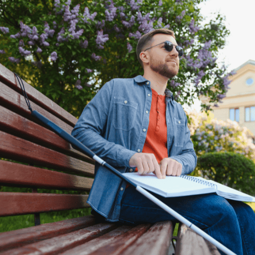 Man with diabetic retinopathy sitting on park bench with cane for the blind reading Louis Braille