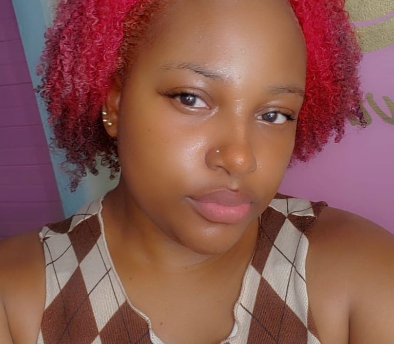 A young woman with rosy pink natural curls, styled in a short, voluminous shape. Her glowing skin, subtle makeup, and nose pi