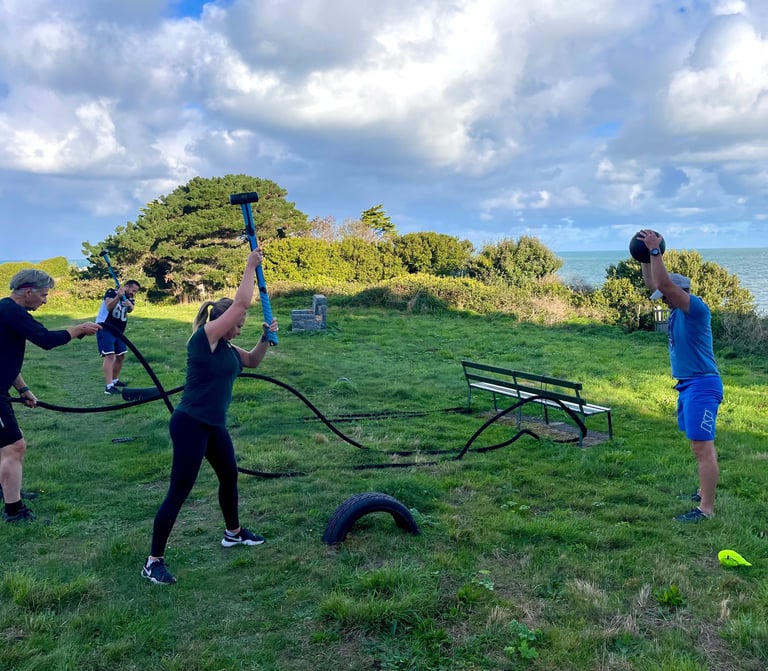 People performing outdoor functional fitness training with battle ropes and slam balls on a coastal lawn.
