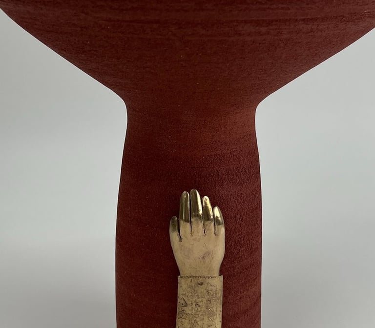 A tall thrown red ceramic vessel, wide at the top. A cast bronze hand is fixed on its base.