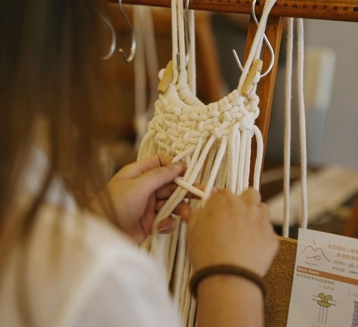 Manos tejiendo pieza de macramé con cordón blanco sobre soporte de madera.