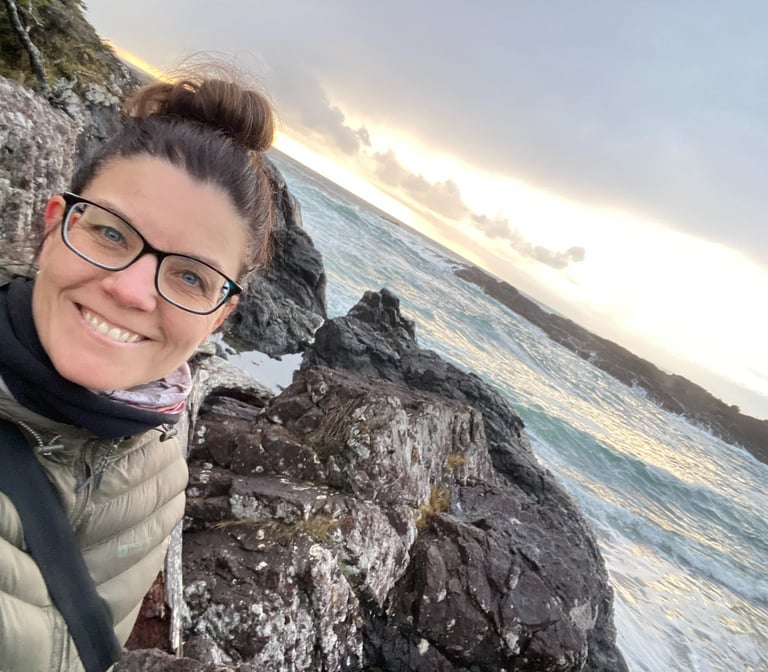 The founder of Alignment Era Consulting smiling on a rocky coast at sunset with ocean waves.