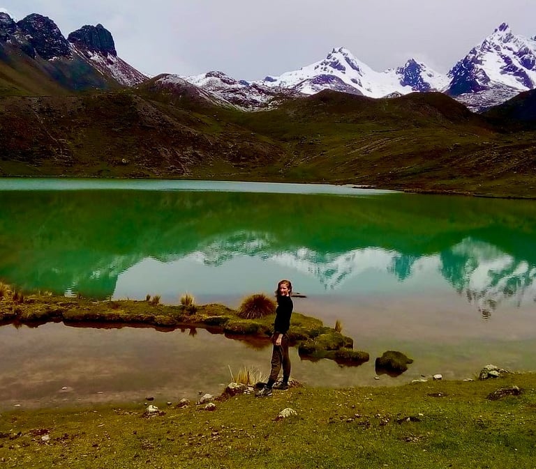A hiker stands by a turquoise lake reflecting snow-capped peaks in the Andes mountains.