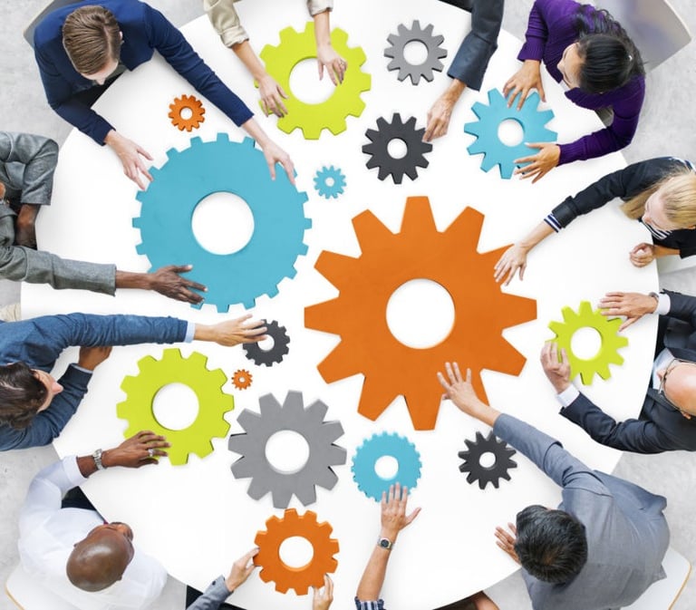 A group of people seated around a table are collaboratively arranging colorful gear-shaped pieces, symbolizing teamwork