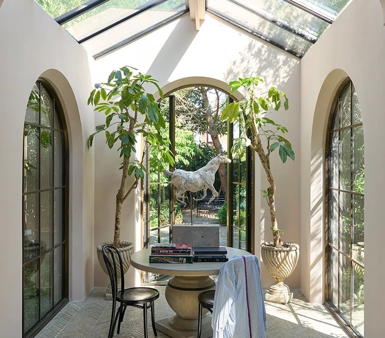 Sunlit garden room with a glass roof, arched windows, indoor trees, and a stone table with a horse sculpture.