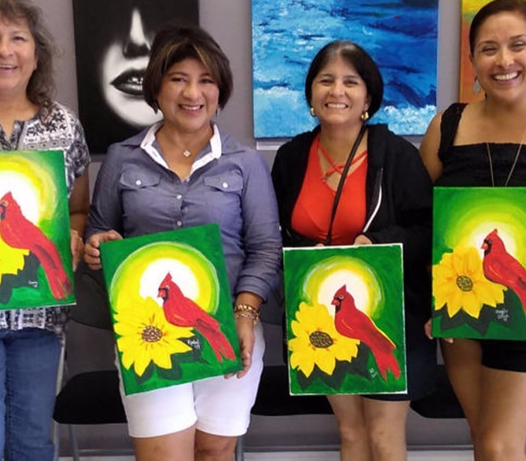 a group of smiling women holding up their paintings of a cardinal and a sunflower