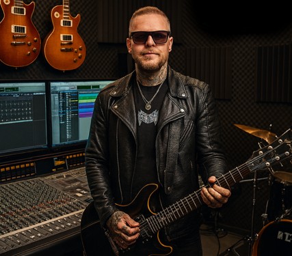 Tattooed music producer in a leather jacket holding an electric guitar in a professional recording studio.