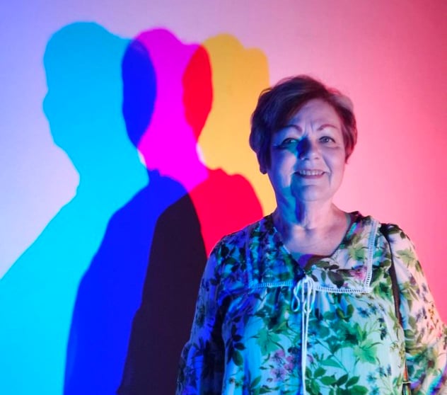 Photo of Debbie with multicolor lights and shadows cast upon the blank wall beside her.