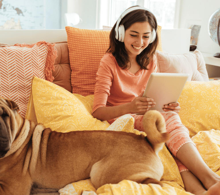 A smiling young girl wearing headphones uses a digital tablet while sitting in bed with her Shar-Pei dog.