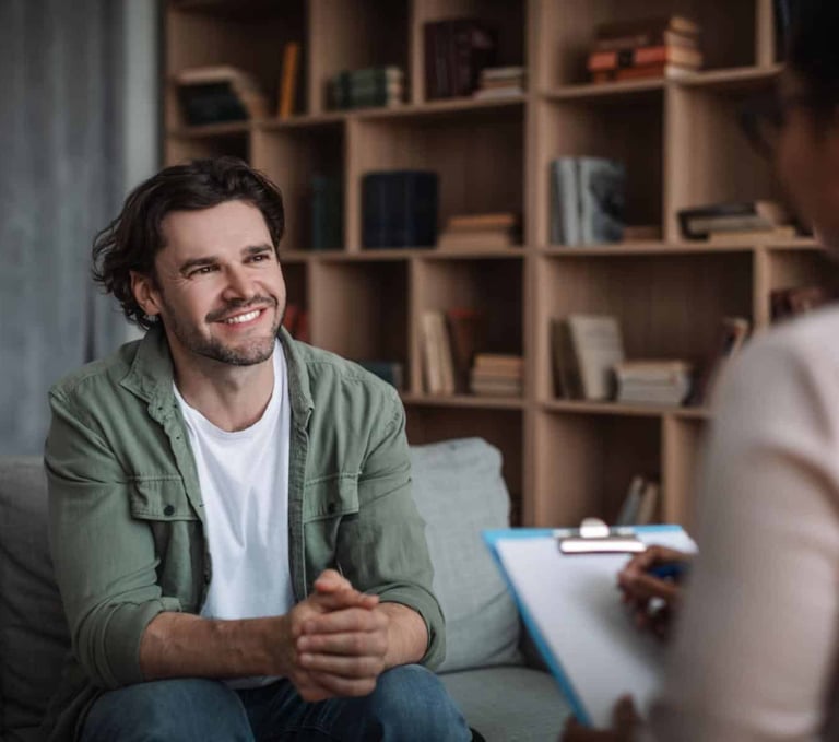 A smiling man sits on a sofa during a mental health therapy session with a counselor.