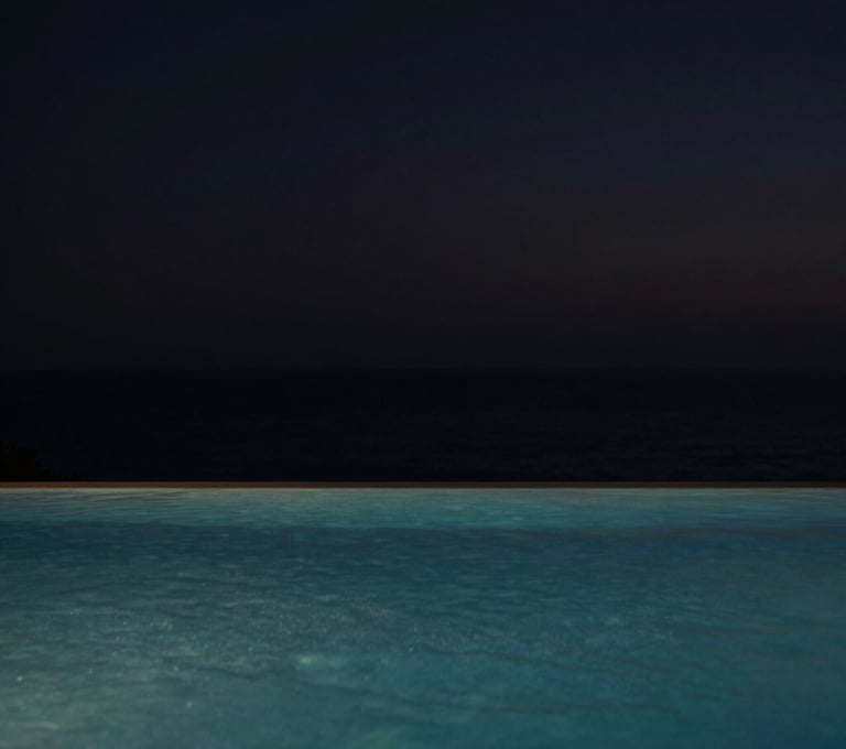 An evocative wide shot of a luxury infinity pool overlooking a coastal horizon at dusk. The water is still, reflecting a deep black sky. Global / Hispano coastal setting. Minimalist, serene, and sophisticated composition.