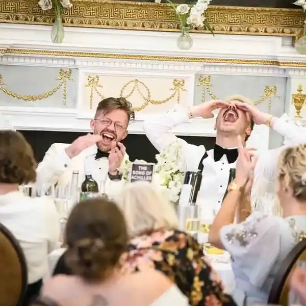 Two grooms in white tuxedos laughing hysterically during wedding reception speeches at a formal head table.