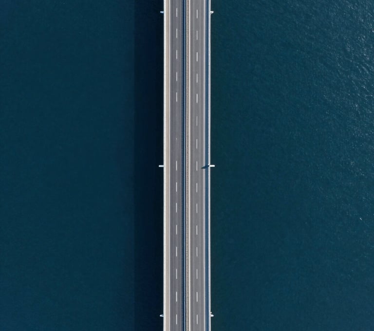 An aerial drone shot looking straight down at a modern coastal highway. The water is deep navy #1B263B and the road is sharp grey #415A77. The composition is symmetrical and high-precision, showcasing high-fidelity drone footage quality.