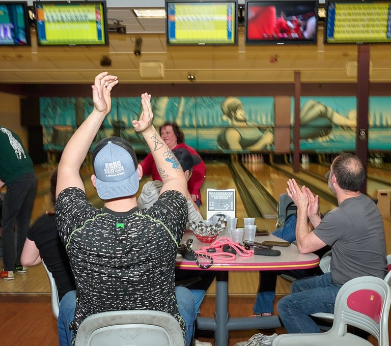 A group of friends celebrating and clapping at a bowling alley with lanes and score monitors.