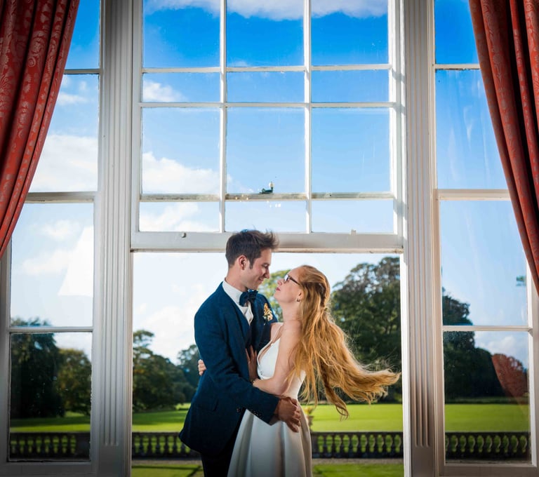 A happy bride and groom embrace in front of a large window overlooking a scenic garden estate.