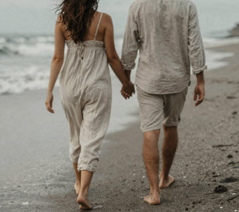 A candid, grain-textured photo of a couple walking barefoot along a quiet Bali beach, their backs to the camera, soft wind blowing through linen clothing, muted and nostalgic #D4C7BB color palette.