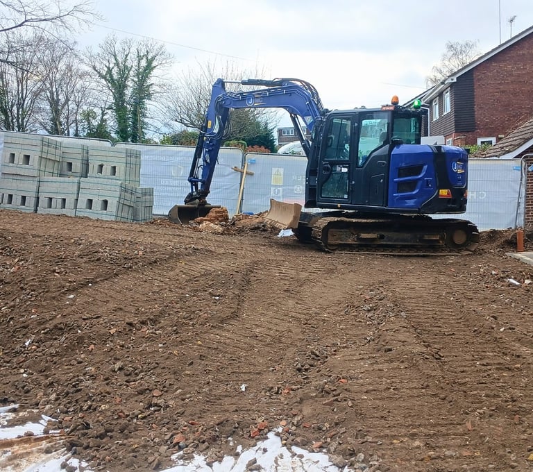 A blue mini excavator and dumper truck on a residential construction site with groundworks in progress.