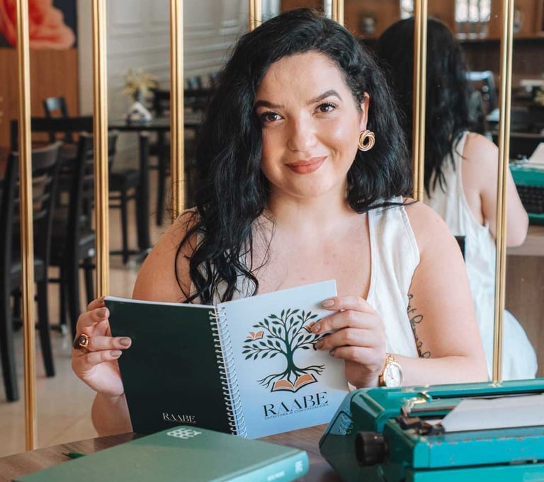 Raabe segurando caderno com livros e máquina de escrever ao lado.