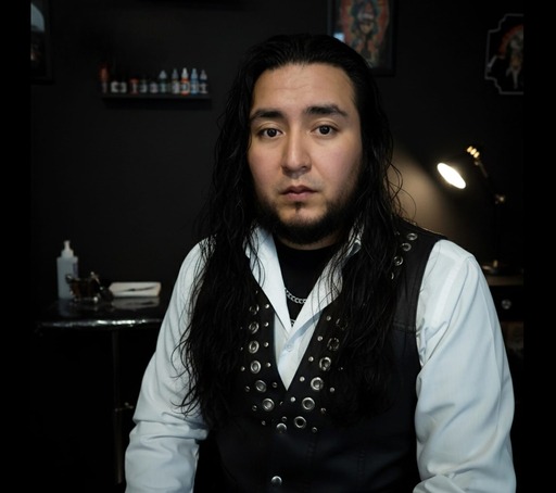 Tatuador profesional masculino con cabello largo y oscuro, usando un chaleco con tachuelas.