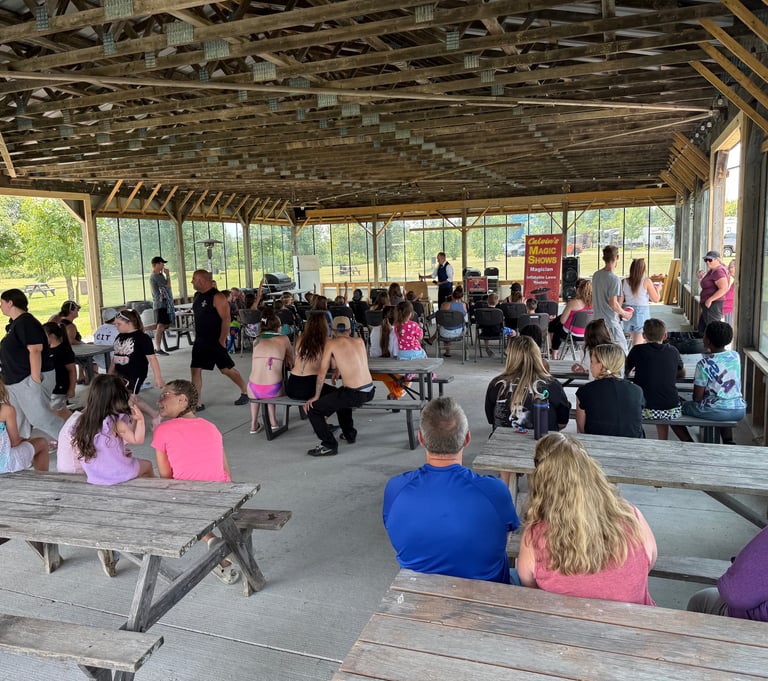 Families gather at picnic tables under a rustic wooden pavilion for an outdoor magic show at a campground.