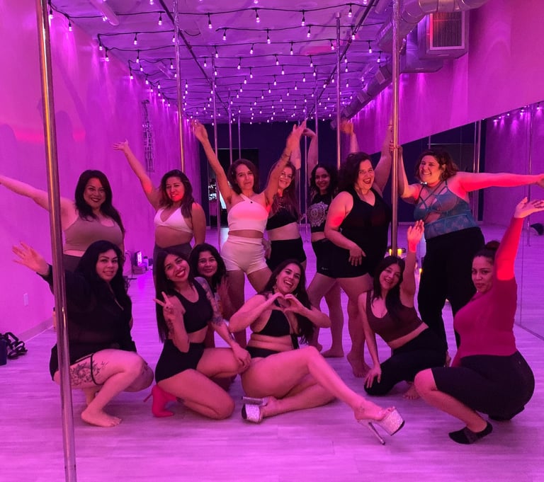 Twelve people posing together in a pole dance studio lit with a pink light