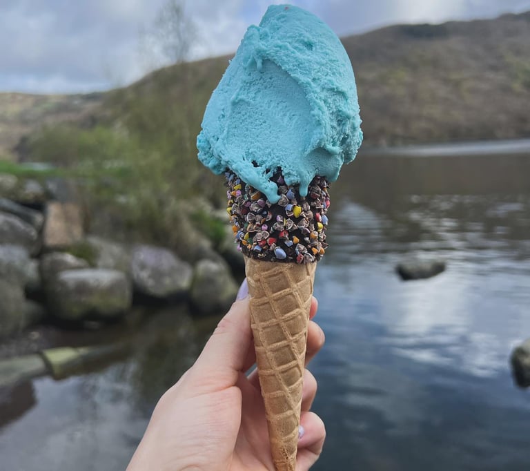Image is a one scoop sprinkle cone with bubblegum ice cream. View of Llyn Padarn in background.
