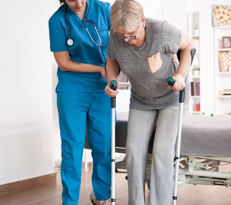 A female nurse in blue scrubs assisting an elderly woman using forearm crutches during physical therapy.