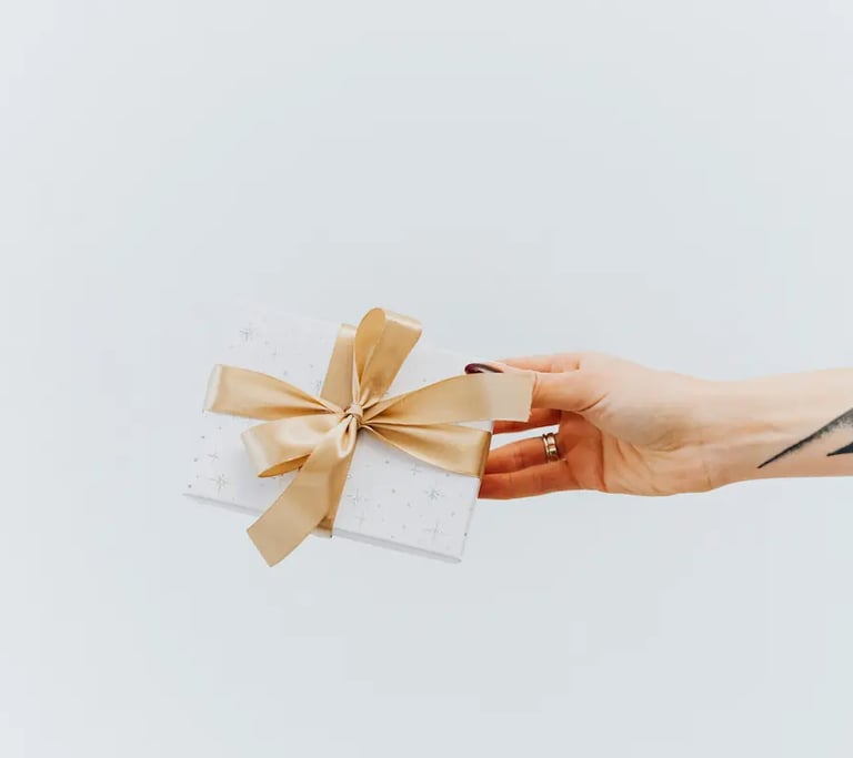 Hand with tattoo holding a white patterned gift box with a gold ribbon bow against a white background.