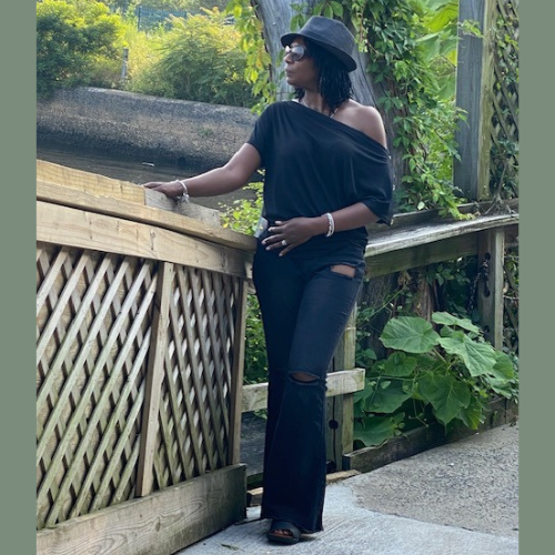 Woman in stylish black off-the-shoulder top and fedora posing on a wooden bridge outdoors.