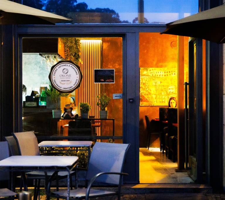 Entrada do Okone Sushi no Porto com mesas externas, fachada de vidro e logótipo iluminado durante o entardecer.