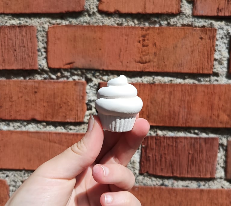 Category tags and tiny objects: Small white ceramic cupcake figurine held against a red brick wall.