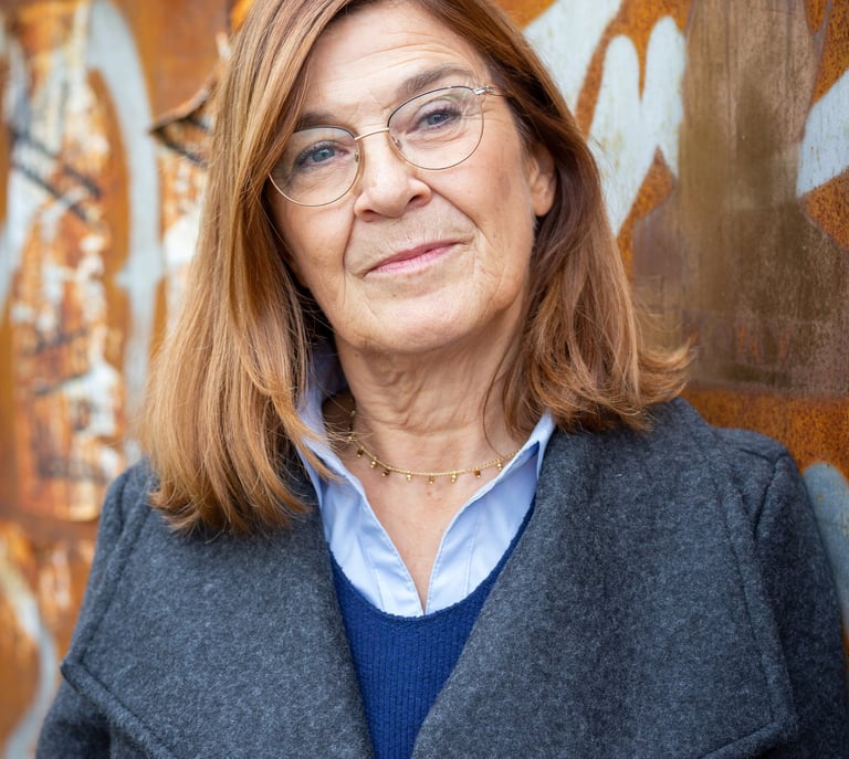 Professional senior woman with glasses and a gray coat standing against a rusty graffiti wall.