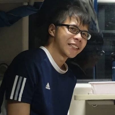 Smiling young Asian man wearing glasses and a blue Adidas t-shirt sitting indoors.