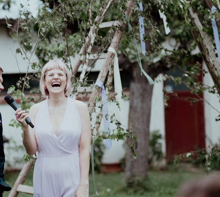 Traurednerin Silke lacht während einer freien Trauung und hält eine persönliche Rede mit Mikrofon im Freien.