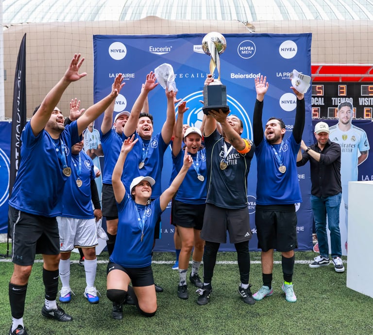 A winning amateur soccer team in blue jerseys celebrates their victory by lifting a silver trophy on a turf field.
