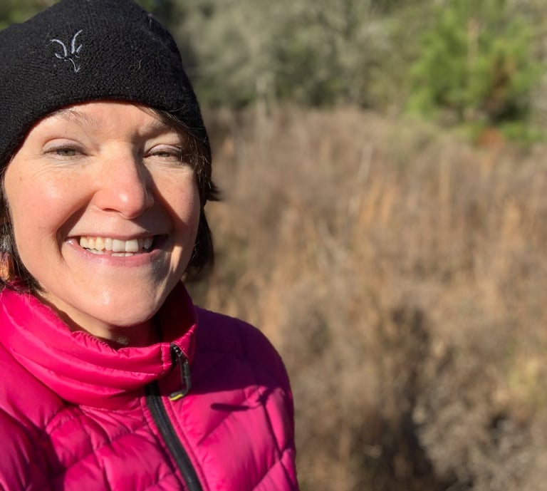 A smiling woman wearing a pink puffer jacket and black beanie hiking outdoors on a sunny day.