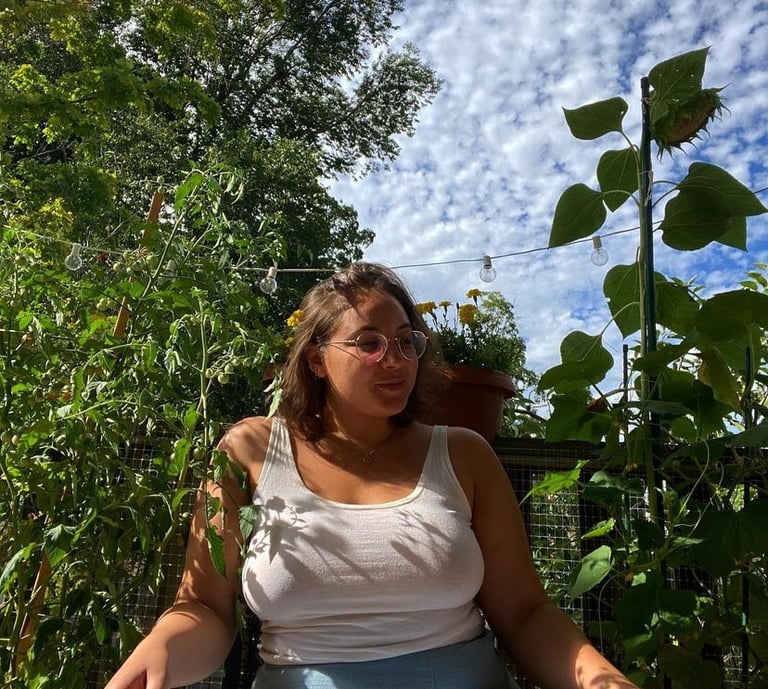 A woman wearing glasses and a white tank top sits in a lush green garden under a blue sky.