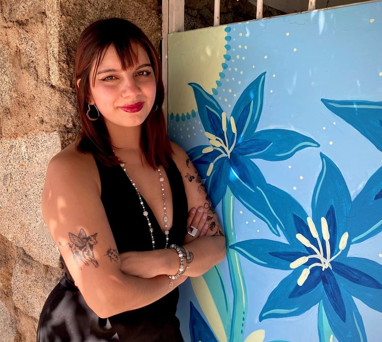 Young woman with tattoos posing in a black dress next to a blue floral mural.